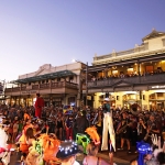 Large crowds enjoying performers playing instruments with stiltwalkers in front of hotel balconies with crowds of people watching