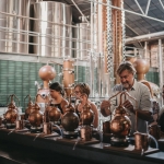 Group of people standing at a collection of miniature stills with large vats behind them in an old warehouse